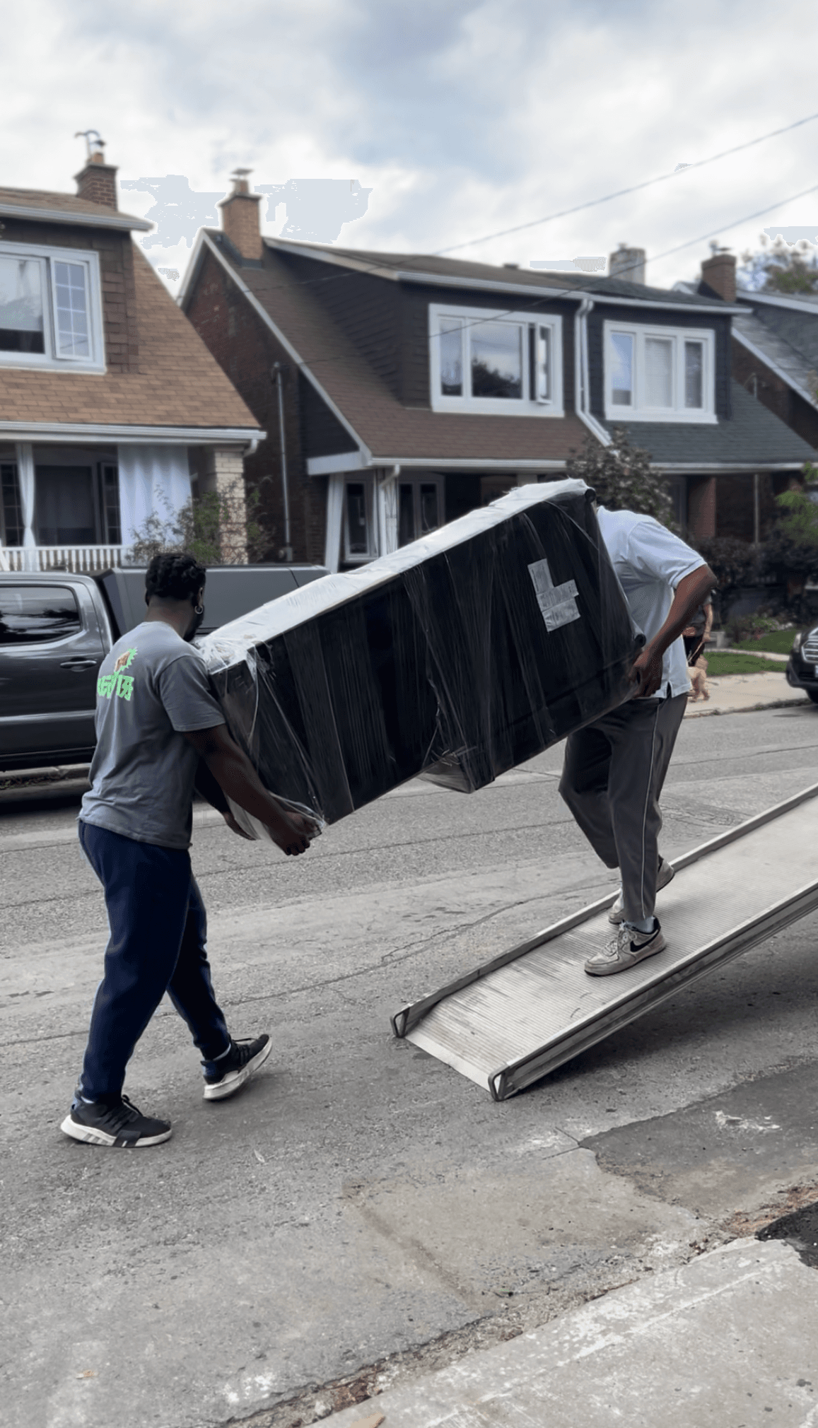Two men carry a large, plastic-wrapped piece of furniture up a metal ramp.